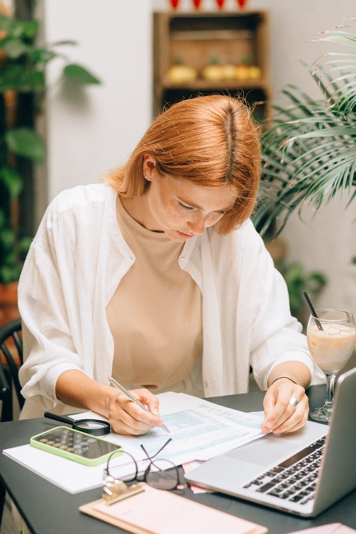 who-we-are Woman analyzing financial documents using laptop and calculator indoors.